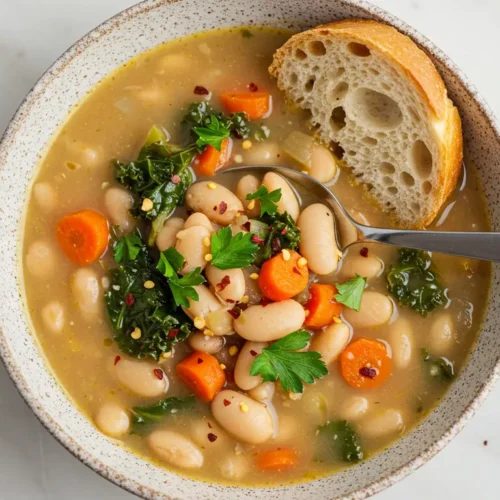 A bowl of Tuscan white bean soup, garnished with fresh parsley and red pepper flakes, accompanied by a slice of crusty bread and a spoon.