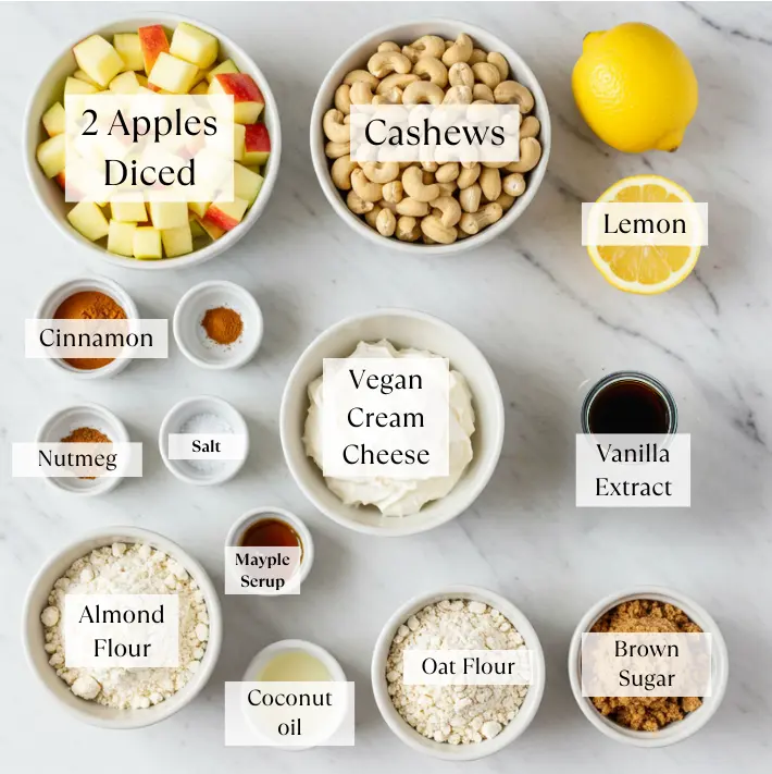 Ingredients for vegan apple crumble cheesecake including fresh apples, oats, cashews, vegan yogurt, spices, lemon, and plant-based butter.